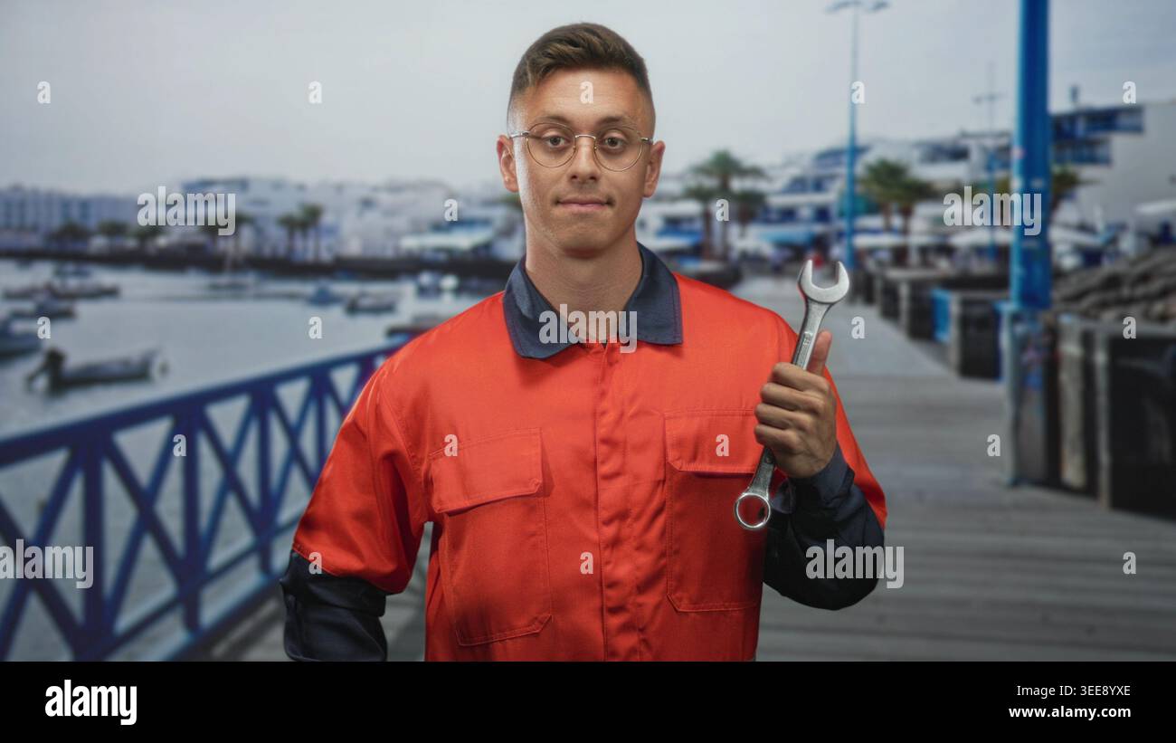 Junger Mechaniker, der einen großen Schraubenschlüssel auf Brusthöhe hält, orangefarbene Overalls und Schutzbrille trägt, während er an einem Pier bei verankerten Booten steht; ruhig Stockfoto