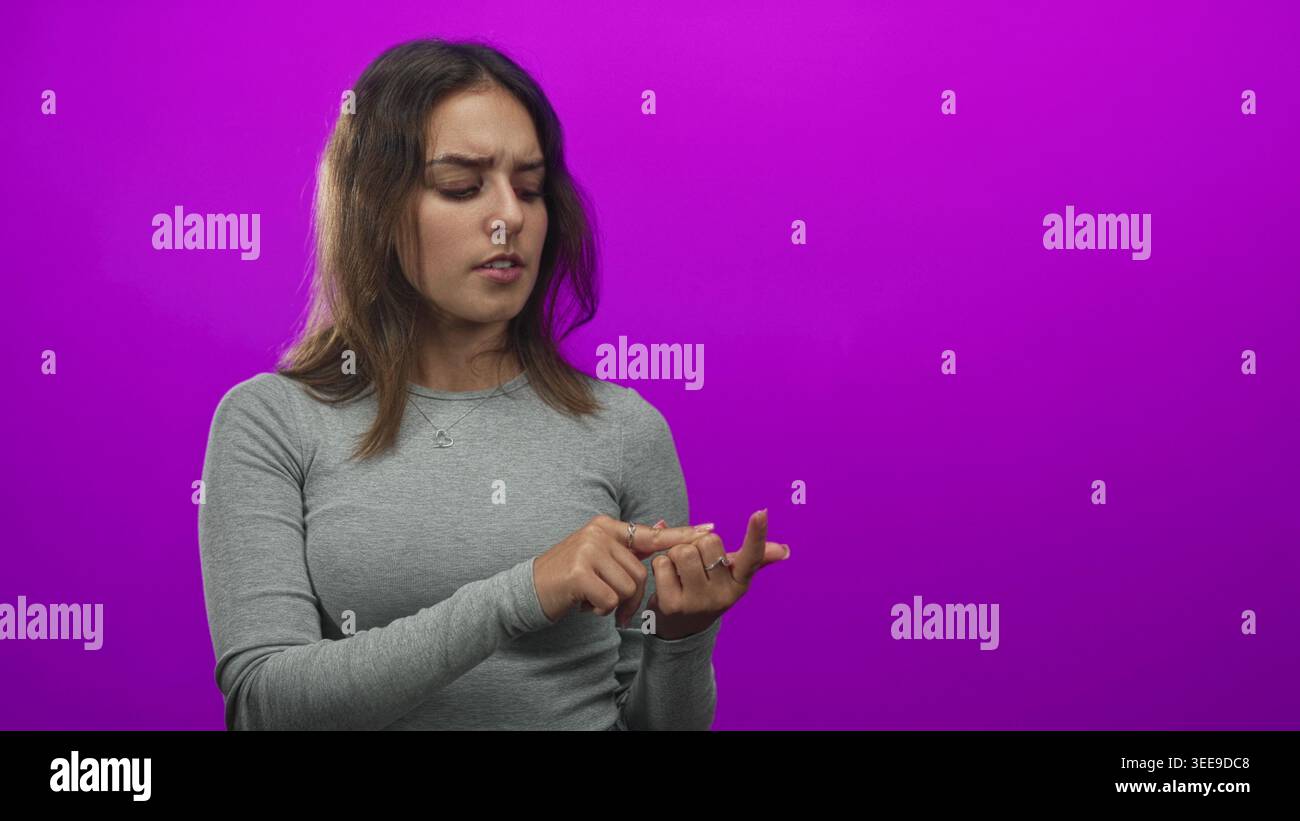 Junge hispanische Frau, die ihre Fingerspitze untersucht, während sie mit der anderen Hand im Studio vor einem violetten Hintergrund zeigt; Sorge. Stockfoto