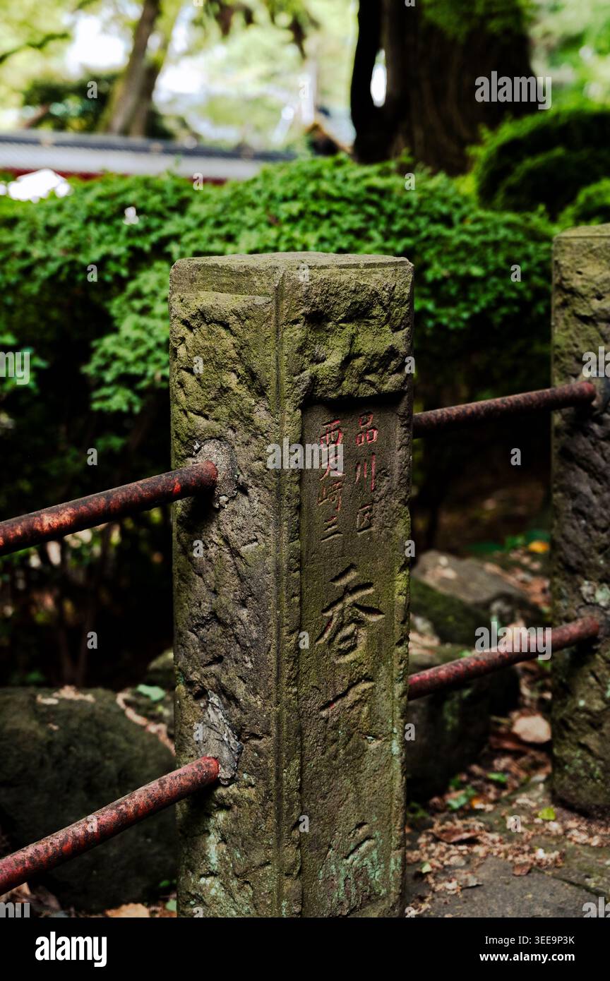 Verwitterter Steinpfosten mit geschnitzter japanischer Inschrift und rostigem Metallgeländer umgeben von Grün am Nezu-Schrein, Bunkyō Bezirk, Tokio, Japan Stockfoto