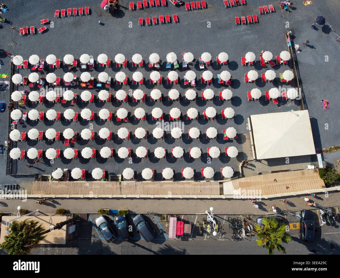 Aus der Vogelperspektive sehen Sie Reihen weißer Sonnenschirme und roter Sonnenliegen an einem dunklen Kieselstrand mit Menschen, die entlang der Promenade in Imperia, Ligu, spazieren gehen und Autos parken Stockfoto