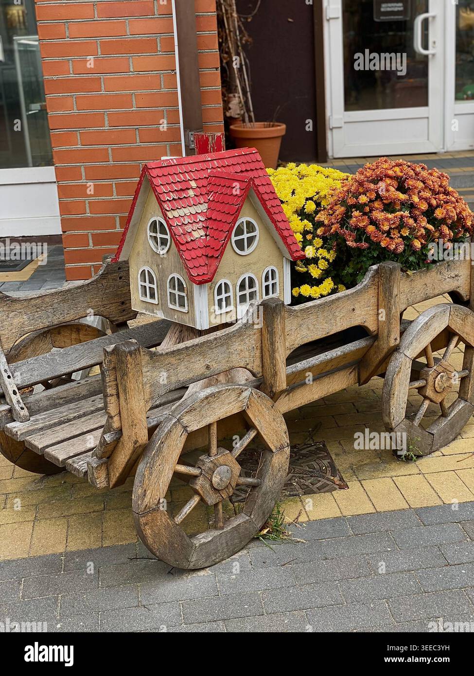 Rustikaler Holzwagen mit dekorativem Miniaturhaus und Herbstblumen in einer gemütlichen Straße - Smartphone-aufgenommenes Stockfoto