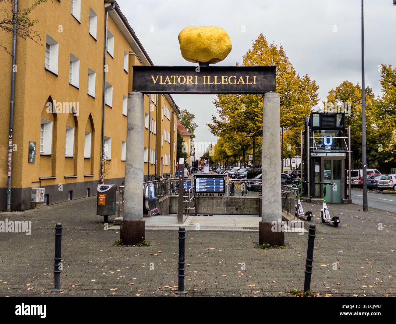 Das Tor mit dem Goldenen Stein markiert den Eingang zur U-Bahn-Station Akazienweg an der Venloer Straße in Köln-Bickendorf, Nordrhein-Westfalen, Deutschland. Die Inschrift "Viatori Illegali" – Latein für "der unrechtmäßige Reisende" – schmückt den Querbalken. Der Kölner Bildhauer Heinrich Brummack schuf das Kunstwerk 1989 im Rahmen eines kommunalen Programms zur Beauftragung von Kunst für die neuen U-Bahn-Stationen in Ehrenfeld. Vor Ort ist es als das "Fahrkarten-Betrüger-Denkmal" bekannt. Herbstfarbene Bäume säumen die Straße im Hintergrund. Die Inschriften spiegeln die laufende Debatte in Deutschland über die Entkriminalisierung von Fahrkarten-Betrug wider Stockfoto