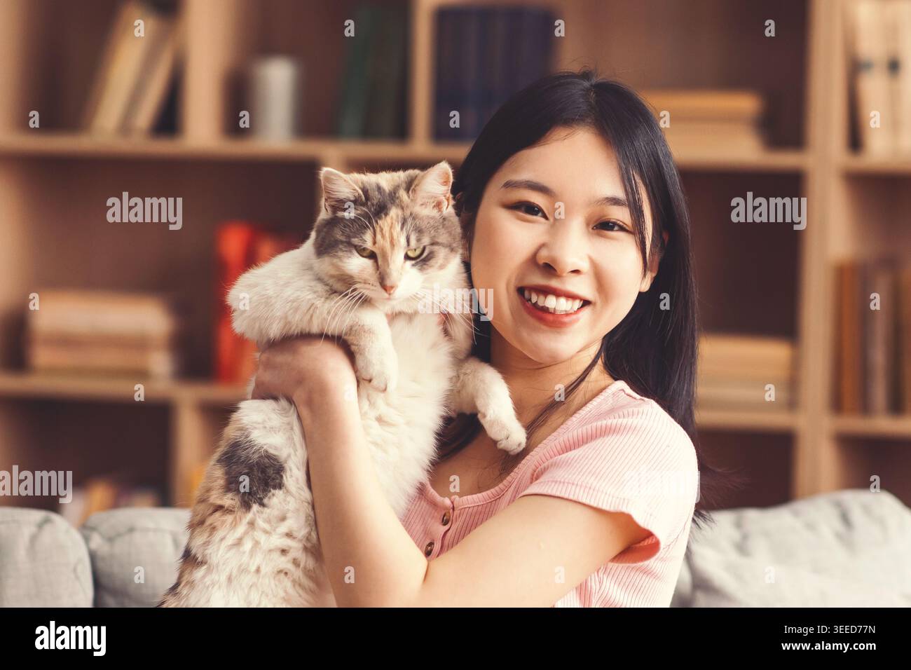 Die junge Frau hält eine Katze und lächelt in einem gemütlichen Innenraum mit Bücherregalen an der Kamera Stockfoto