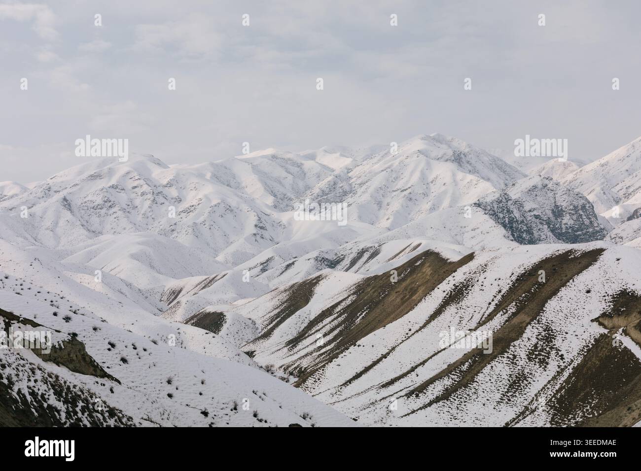 Panoramablick auf schneebedeckte Bergkämme und Gipfel in Kirgisistan, Zentralasien, mit mehrschichtigen Hängen, felsigen Strukturen und einem bewölkten Winterhimmel Stockfoto