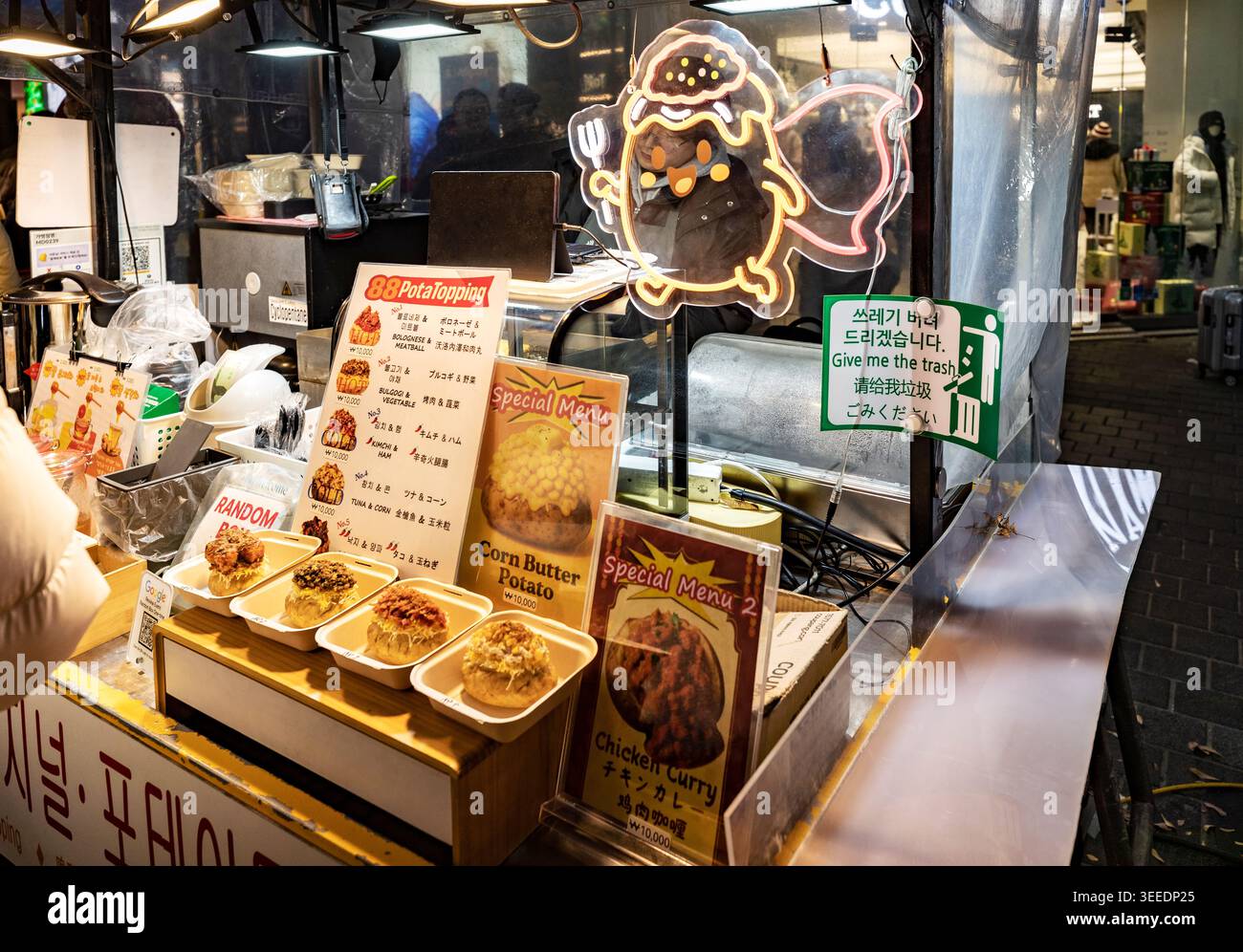 Ein beladener gegrillter Kartoffelstand zeigt Maisbutter und Hühnercurry-Toppings unter einem Neon-Maskottchen-Schild am Myeongdong-Nachtmarkt in Seoul, Südkorea Stockfoto