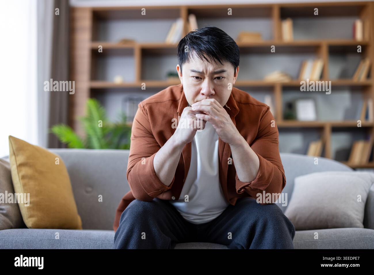 Asiatischer Mann, der sich tief auf ein Problem konzentriert, Stress oder Angst erlebt, während er auf einer Couch in seinem modernen Wohnzimmer sitzt und über Einsamkeit oder psychische Gesundheit reflektiert Stockfoto