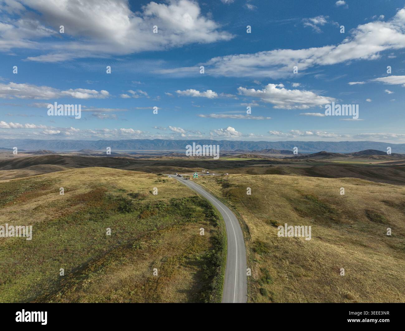 Aus der Vogelperspektive ist eine lange, kurvenreiche Asphaltstraße zu sehen, die über offene, sanfte Hügel mit trockenem, goldbraunem Pflanzenwuchs unter einem breiten Kreis verläuft Stockfoto