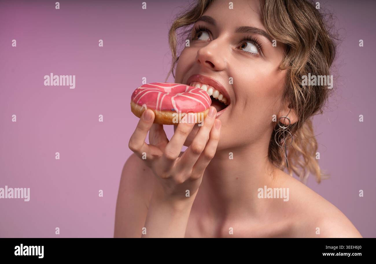 Eine junge Frau isst einen rosa glasierten Donut, genießt ein süßes Dessert, sehnt sich nach mehr zuckerhaltigen Leckereien und genießt einen Snack Stockfoto
