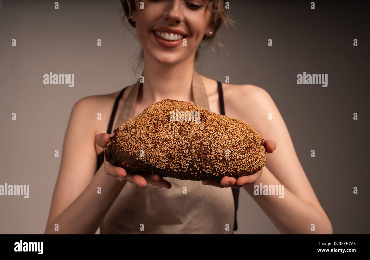 Junge Bäckerin, die ein frisch gebackenes dunkles Brot mit Sesamsamen hält und präsentiert, lächelnd Stockfoto