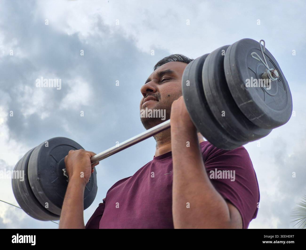 Ein 30-jähriger indischer Mann, der Barbell mit geschlossenen Augen hebt, Krafttraining und Muskelaufbau im mittleren Alter, gesunder Lebensstil, Mann Workout. - Smartphone-aufgenommenes Stockfoto