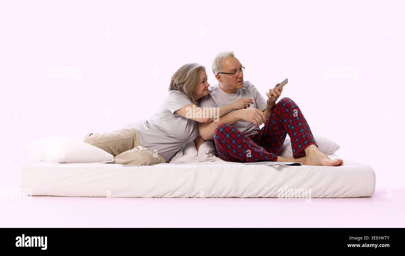 Seniorenpaar, das zusammen ein Smartphone benutzt, während sie auf dem Bett vor weichem rosa Hintergrund sitzt Stockfoto