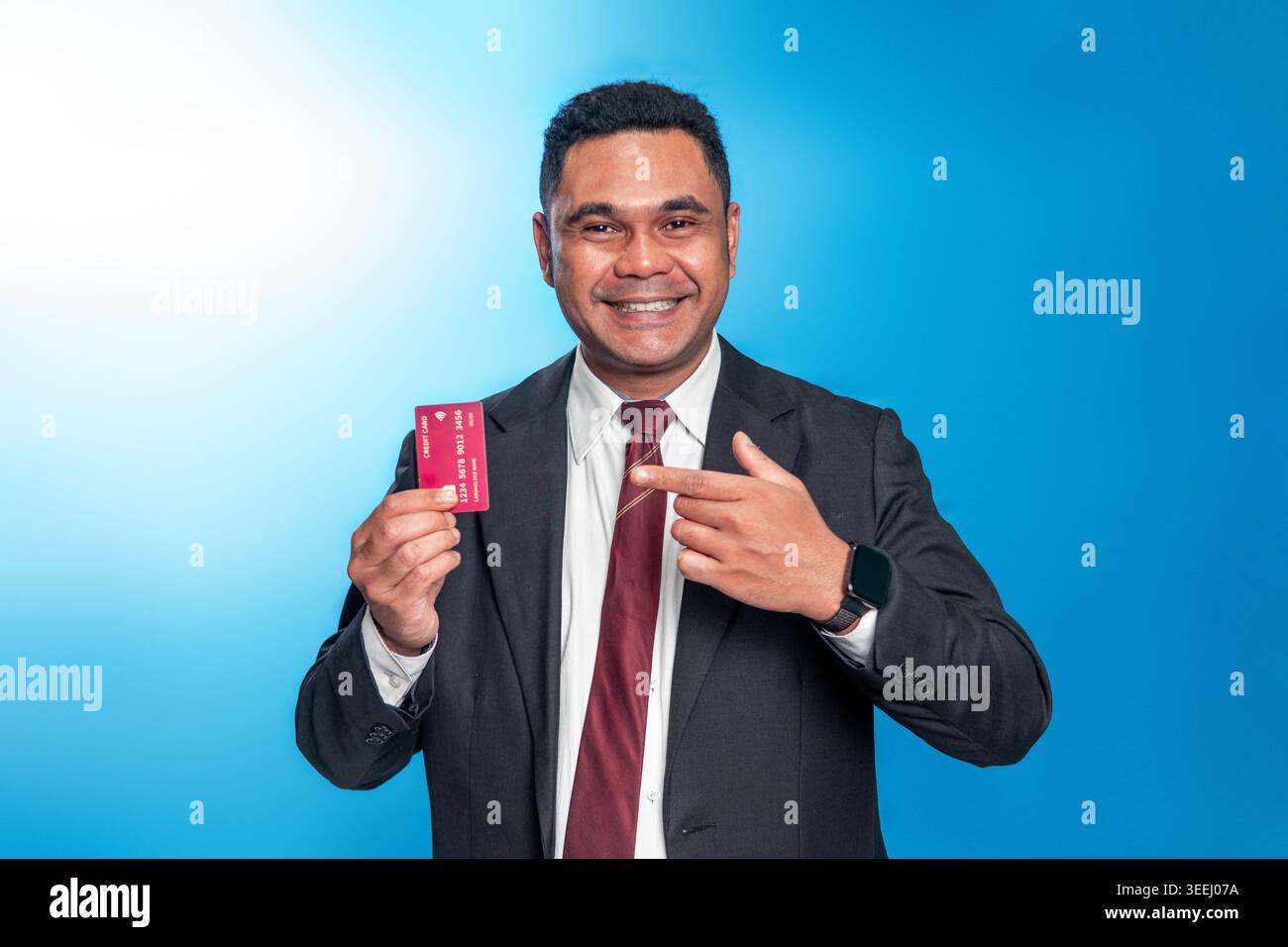 Lächelnder indonesischer Mann in schwarzem Anzug und Krawatte, der eine Kreditkarte hält und darauf zeigt. Studio-Aufnahme auf blauem Hintergrund für Finanz- und Bankwesen. Stockfoto