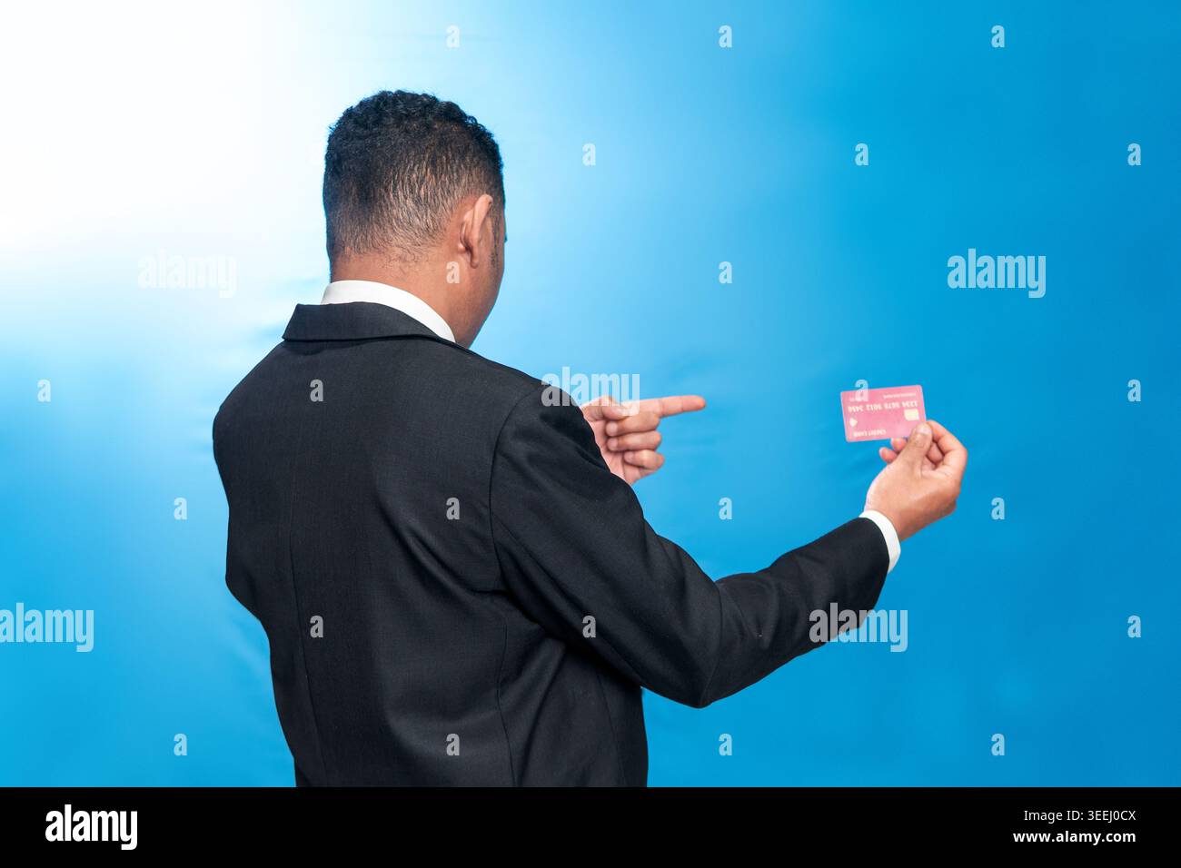 Rückansicht eines indonesischen Mannes in einem Anzug, der eine rote Kreditkarte vor blauem Hintergrund hält. Konzept des finanziellen Erfolgs und des Premium-Zugangs. Stockfoto