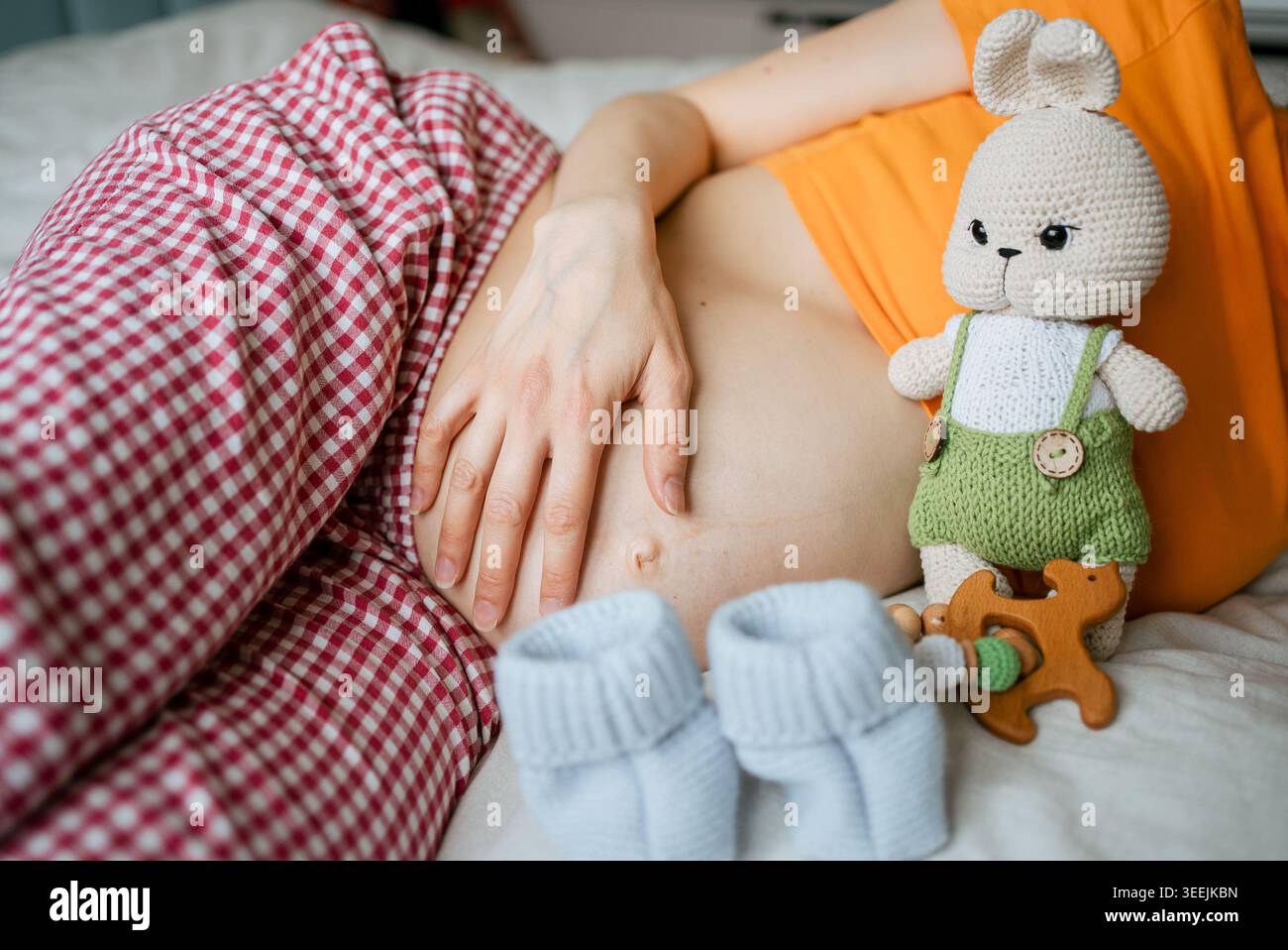 Schwangere Frau, die auf dem Bett liegt und sanft ihren Babybummel berührt, umgeben von Babyartikeln wie einem gestrickten Häschen, Stiefeln und einem hölzernen Beißring, die die bevorstehende Mutterschaft und den Neuanfang repräsentieren Stockfoto