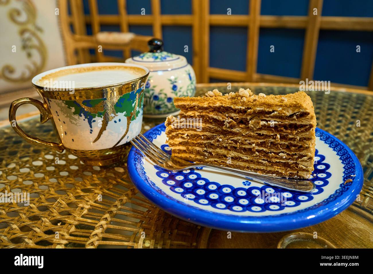 Ein Stück Honig-Kuchen auf einer blauen Keramikplatte mit Gabel, serviert mit einer Tasse Cappuccino-Kaffee in einem gemütlichen Café. Stockfoto