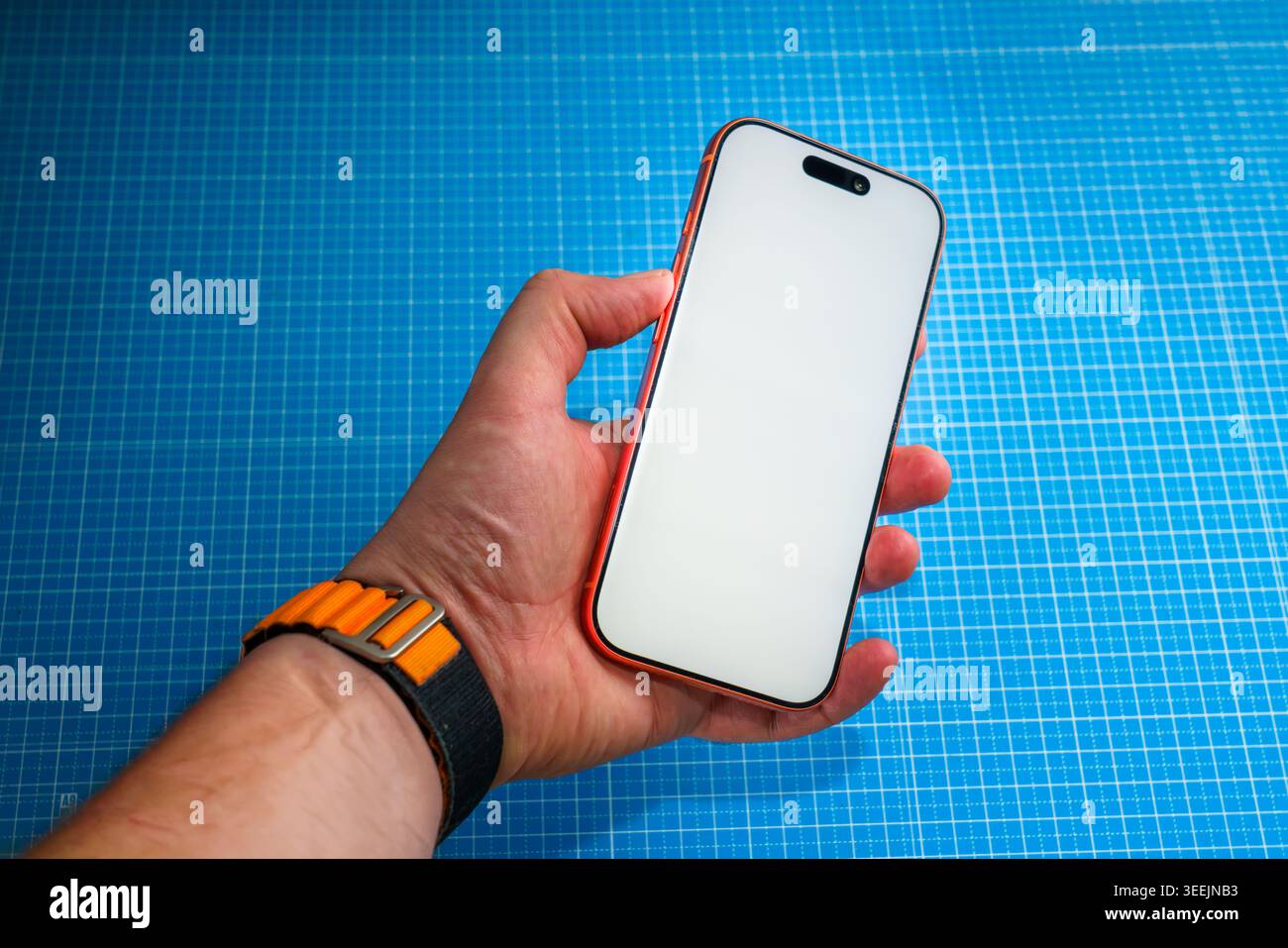 POSEN, POLEN - 11. MAI 2026: Die Hand eines Mannes hält ein oranges iPhone mit einem leeren weißen Bildschirm und Dynamic Island vor einem blauen Gitterhintergrund, Tragen Stockfoto