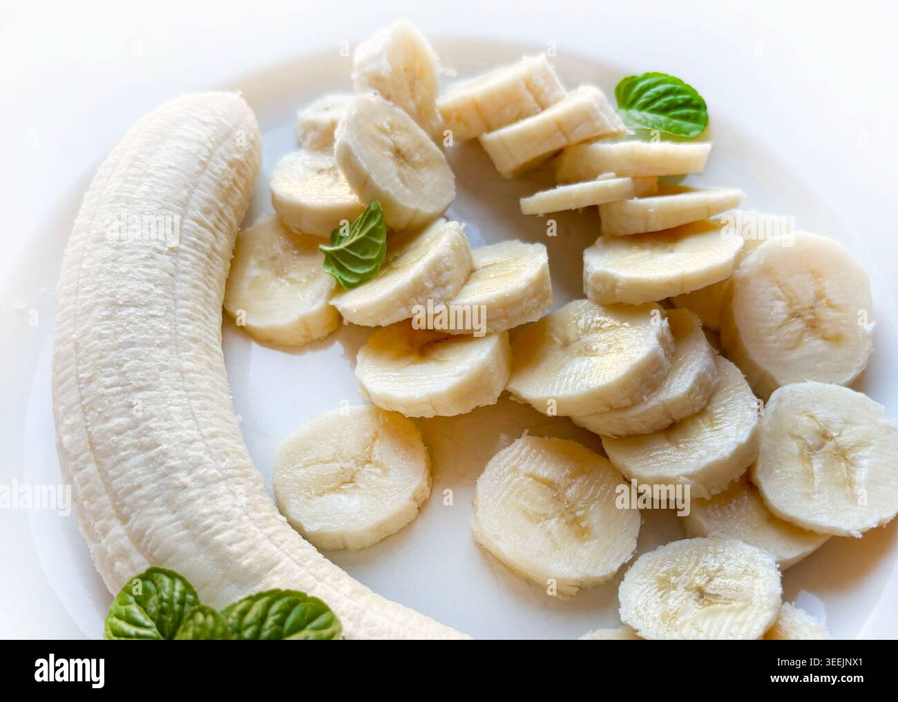 Blick von oben auf eine ganze geschälte Banane und frische Scheiben auf weißem Hintergrund, dekoriert mit grünen Minzblättern. Stockfoto