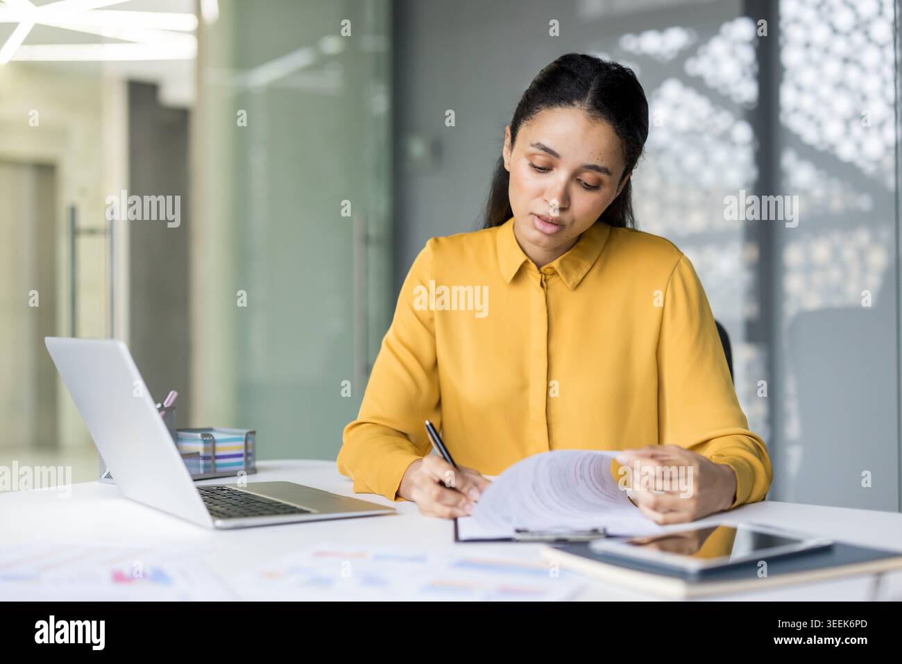 Junge professionelle Frau, die Dokumente überprüft und Notizen an ihrem Schreibtisch macht, zeigt Konzentration und Hingabe für Aufgaben in einer modernen Büroumgebung Stockfoto
