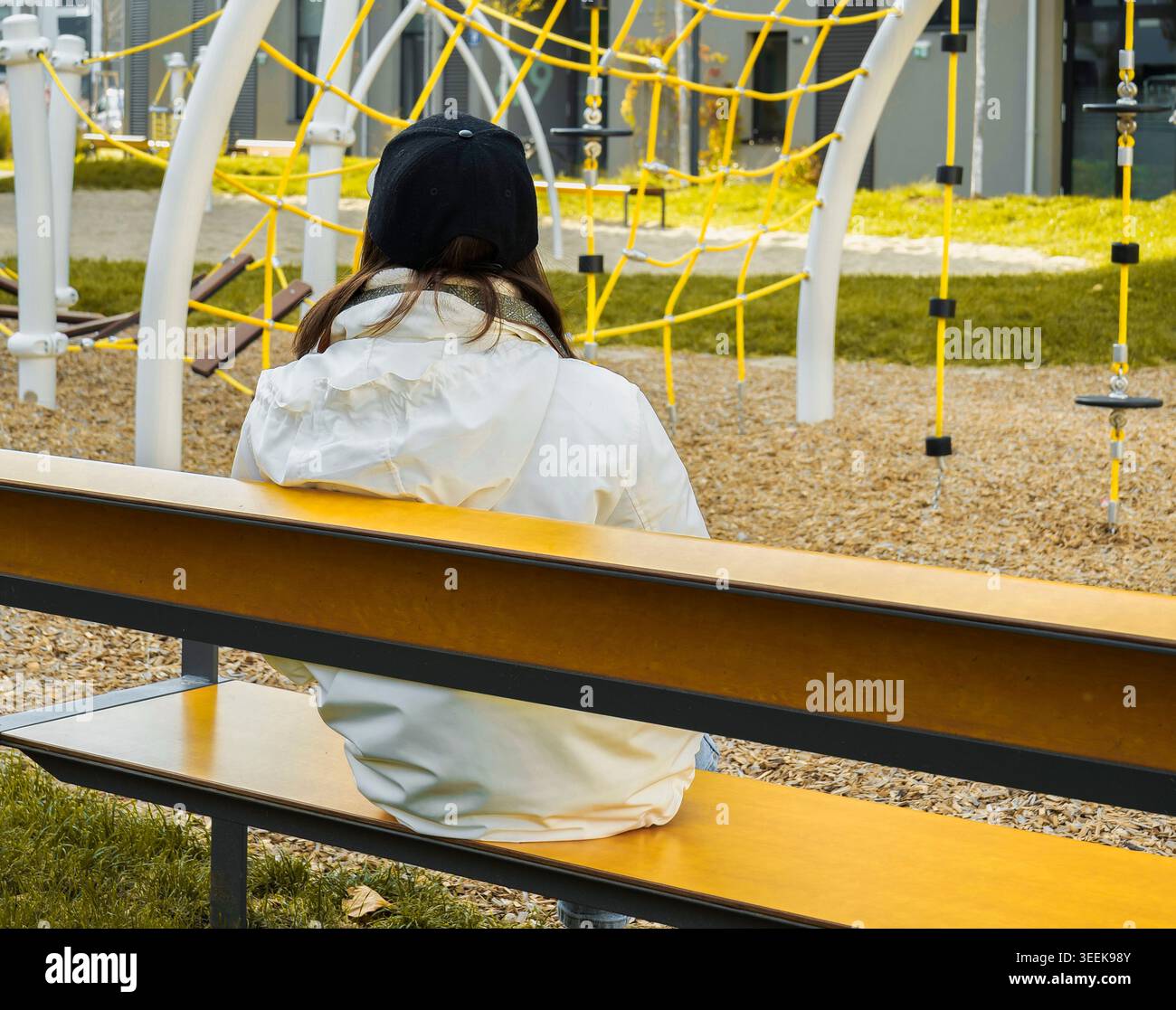 Die junge Frau in einer weißen Jacke sitzt leise auf einer Bank und blickt auf einen modernen Spielplatz mit heller Kletterausrüstung Stockfoto