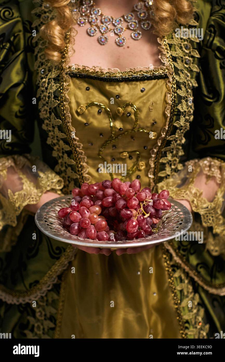 Vertikale Aufnahme einer Frau, die einen Teller mit roten Trauben hält, während sie ein historisches Kleid trägt und ein luxuriöses Lebensmittelkonzept und ein Vintage-Gastfreundschaft-Thema für das Event-Marketing präsentiert Stockfoto