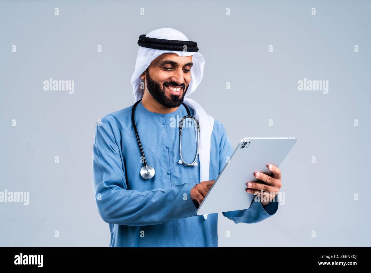 Mann aus dem Nahen Osten in traditioneller emiratischer Kleidung und einem Stethoskop, lächelnd, während er ein digitales Tablet benutzt, repräsentiert moderne Gesundheitsfürsorge, Arzt Stockfoto
