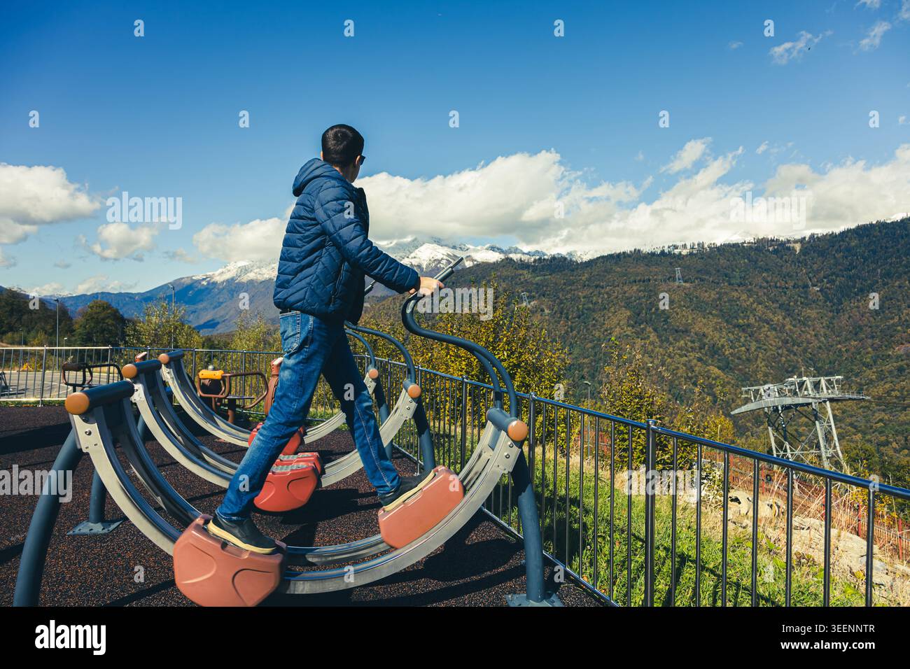Ein Junge steht auf Fitnessgeräten in einem Fitnessbereich im Freien. Er sieht schneebedeckte Berge unter blauem Himmel. Im Fitnessbereich sind Bäume zu sehen. Stockfoto