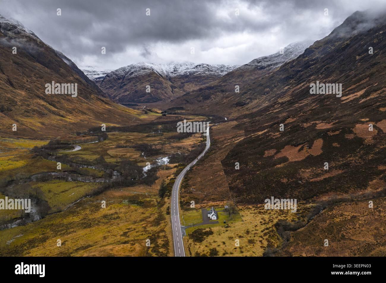 Aus der Vogelperspektive auf die gewundene A82-Straße durch das dramatische Glencoe-Tal mit schneebedeckten Bergen und einem kleinen weißen Häuschen unter einem stimmungsvollen Himmel Scotlan Stockfoto