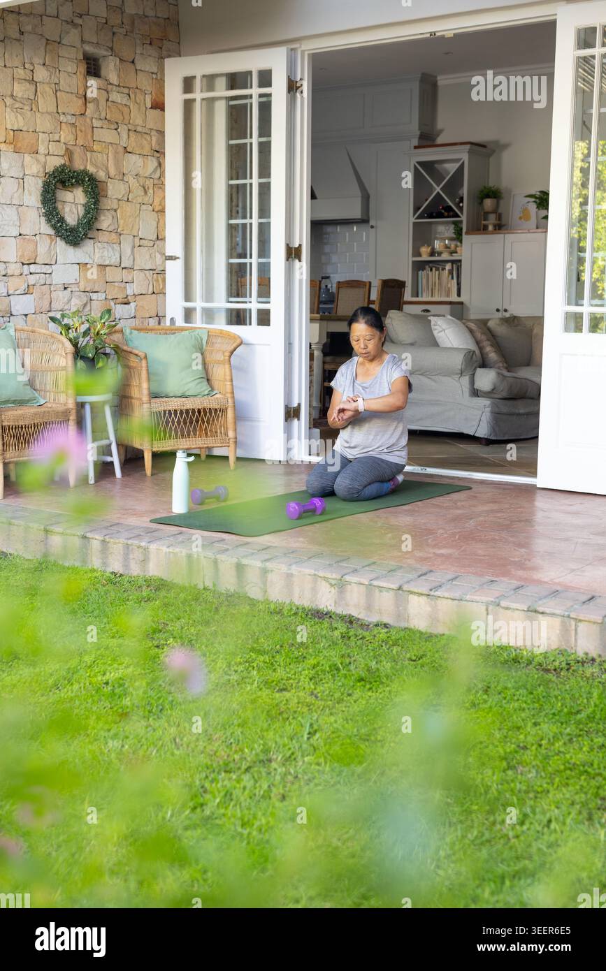 Ältere asiatische Frau kniend auf grüner Terrassenmatte, um Uhr in Top dunklen Leggings in der Nähe von Kurzhanteln zu sehen Stockfoto