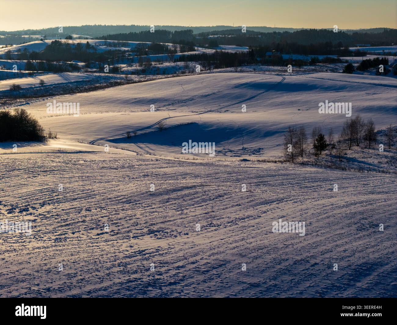 Aus der Vogelperspektive auf schneebedeckte Hügel und Felder mit langen Schatten und Pfaden während der goldenen Stunde in Litauen. Stockfoto