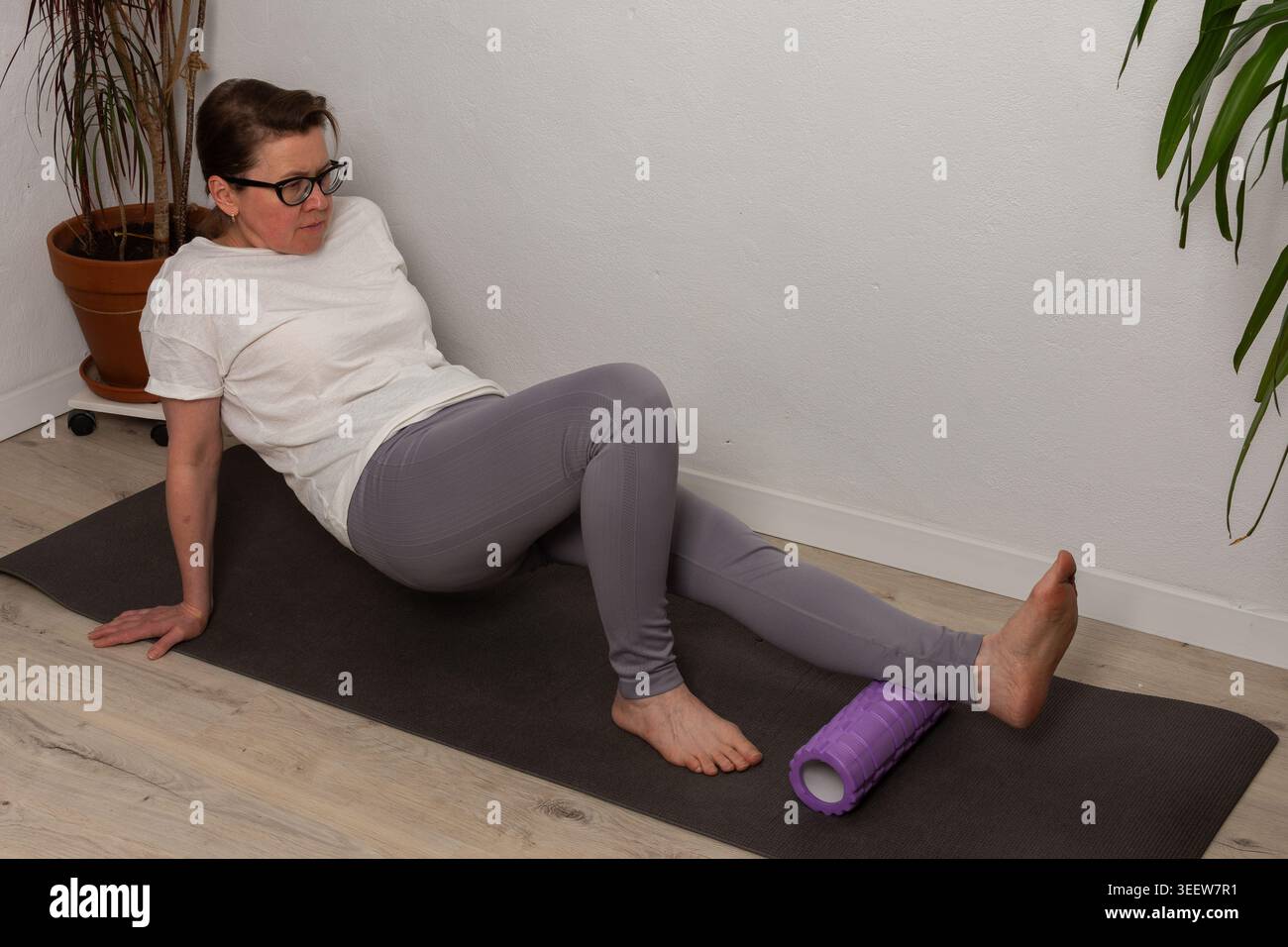 Eine 52-jährige Frau übt zu Hause auf einer Matte. Sie verwendet eine Schaumstoffrolle zum Dehnen und Genesen, während sie auf dem Boden sitzt. Pflanzen sind während ihres Trainings im Hintergrund Stockfoto