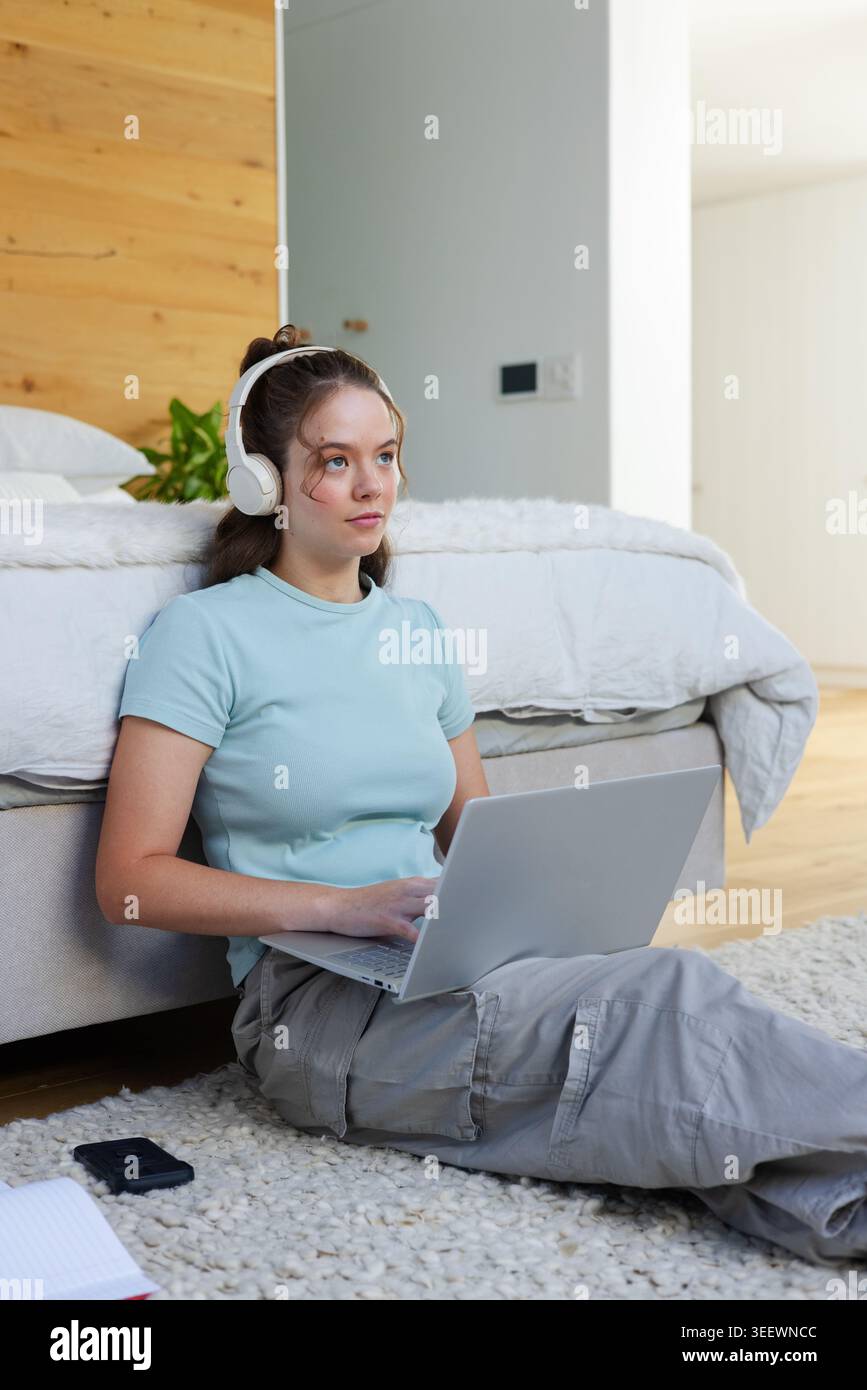 Frau, die auf zotteligem Schlafzimmerteppich sitzt, Kopfhörer und hellblaues Oberteil trägt und auf dem Laptop schreibt Stockfoto