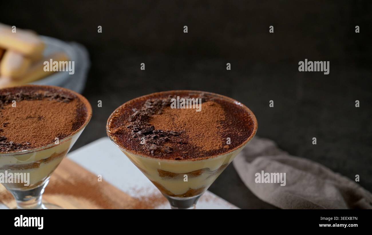 Einzelne Portionen cremigem Tiramisu werden in eleganten Cocktailgläsern mit Stielstiel vor dunklem Hintergrund serviert. Das Dessert bietet sichtbare Speisen Stockfoto