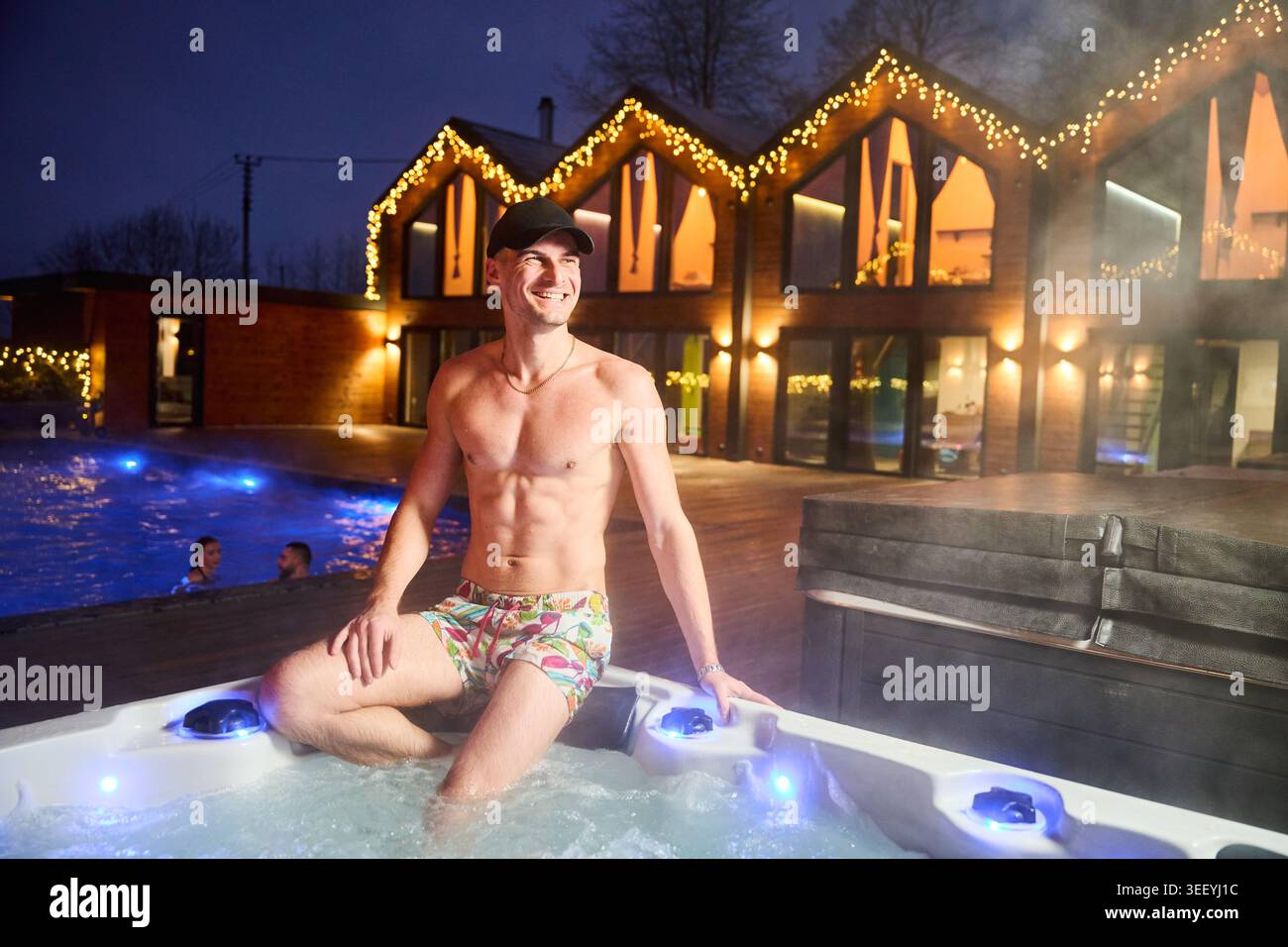 Mann, der sich nachts im Whirlpool im Freien in der Nähe eines modernen, beleuchteten Hauses entspannt, das luxuriöse Lifestyle, Wellness, Freizeit und ein erstklassiges Urlaubserlebnis repräsentiert Stockfoto