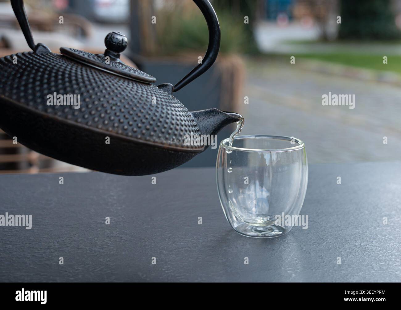 Grüner Tee aus einem Metallkessel auf verschwommenem Hintergrund in ein Glas gießen, heißes Getränk in der Teekanne im japanischen Restaurant, dunkle Tischplatte Stockfoto