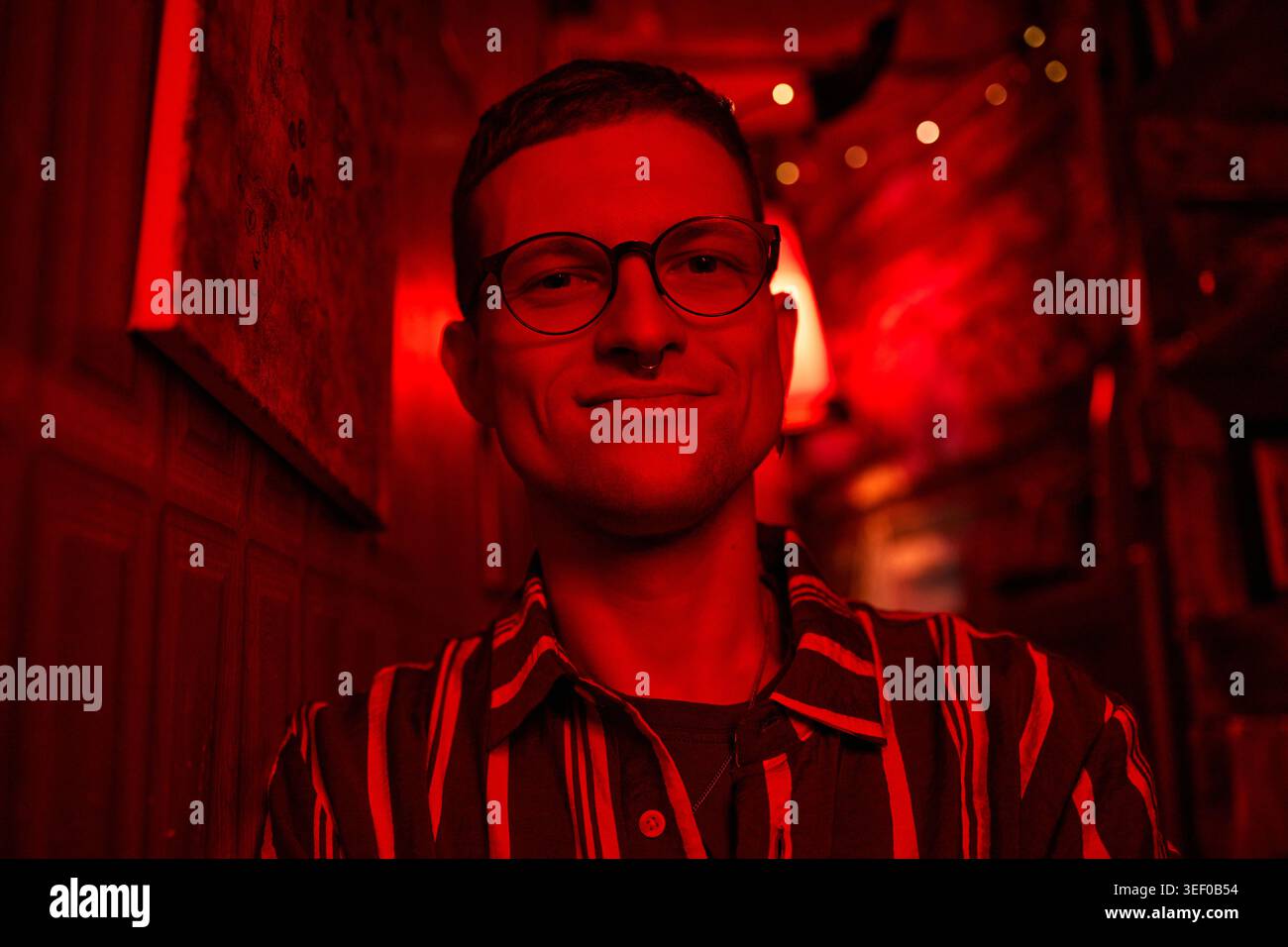 Porträt eines jungen Erwachsenen kaukasischen Mannes mit Brille, Blick auf die Kamera und Lächeln im Nachtleben, was auf Selbstvertrauen und Identität hindeutet. Nützlich für Dating-Apps, soziale Kampagnen, Lifestyle-Branding Stockfoto