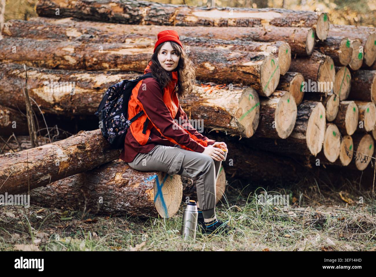 Die Frau sitzt auf gestapelten Baumstämmen im Wald und hält eine Tasse mit Thermoskanne auf dem Boden. Überlebensbereitschaft im Freien, warm in wilder Wildnis Stockfoto