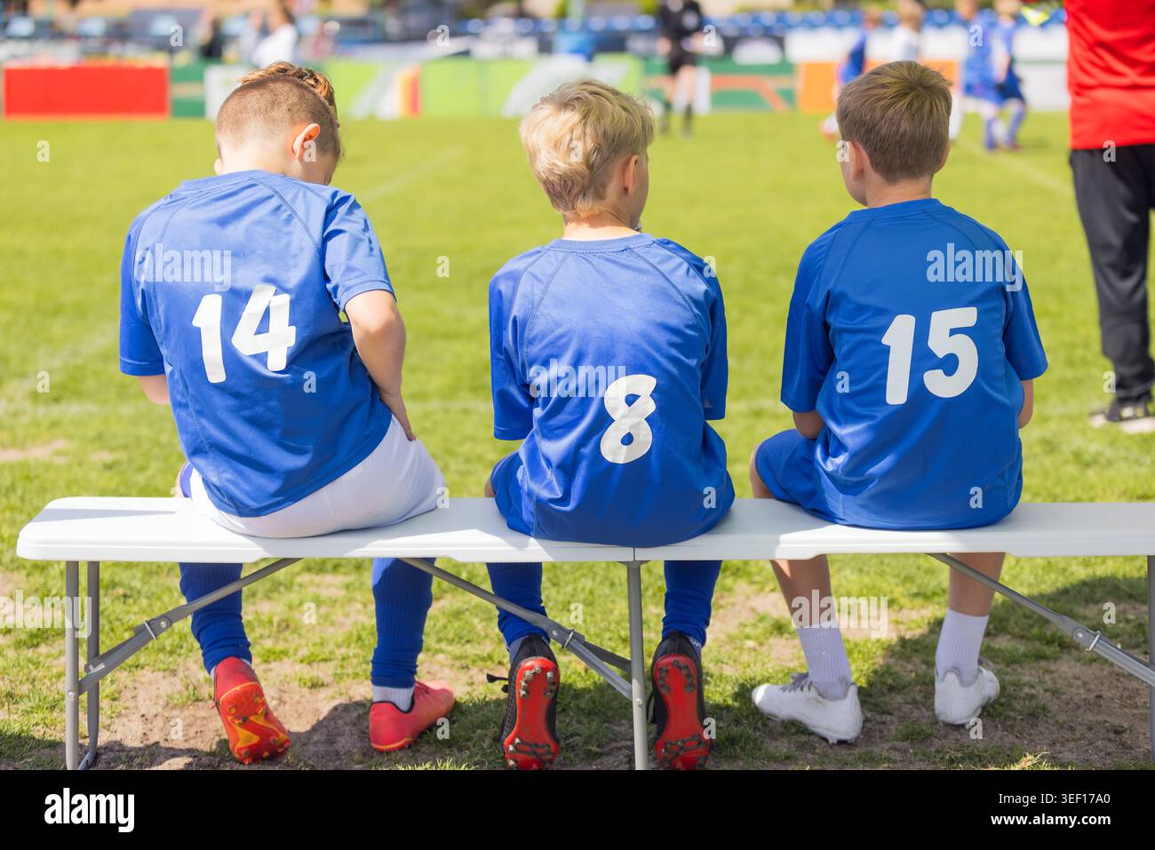 Junior Soccer Substituts in blau nummerierten Trikots sitzen auf der Sideline Bank und schauen sich das Fußballspiel an – Team Management und Spielerentwicklung Stockfoto