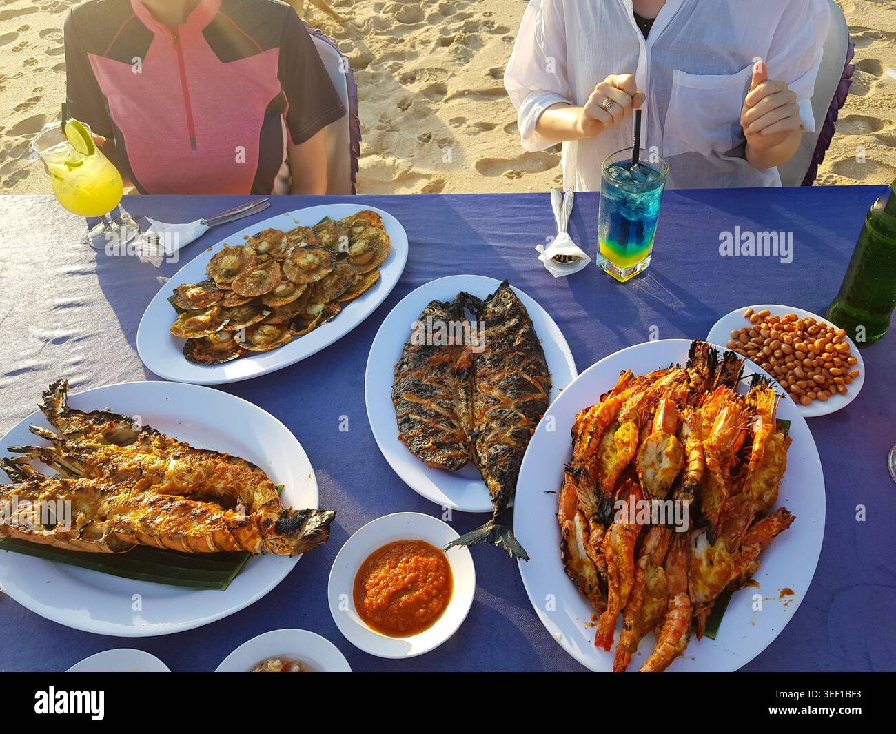 Frisch gegrillte Meeresfrüchte, darunter Garnelen, Fisch und Meeresfrüchte, werden während des Sonnenuntergangs an einem Strandtisch serviert. Tropische Urlaubs-Dining Stockfoto
