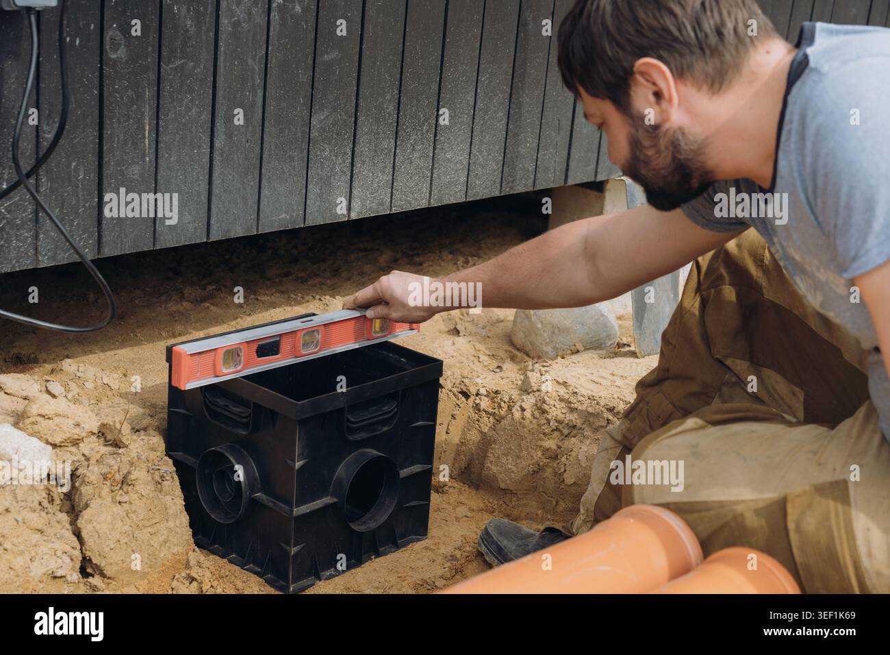 Ein Mann installiert ein Regenwasser-Auffangbecken für das Entwässerungssystem eines Landhauses und überprüft seine Position mit einer Wasserwaage. Hochwertige Fotos Stockfoto