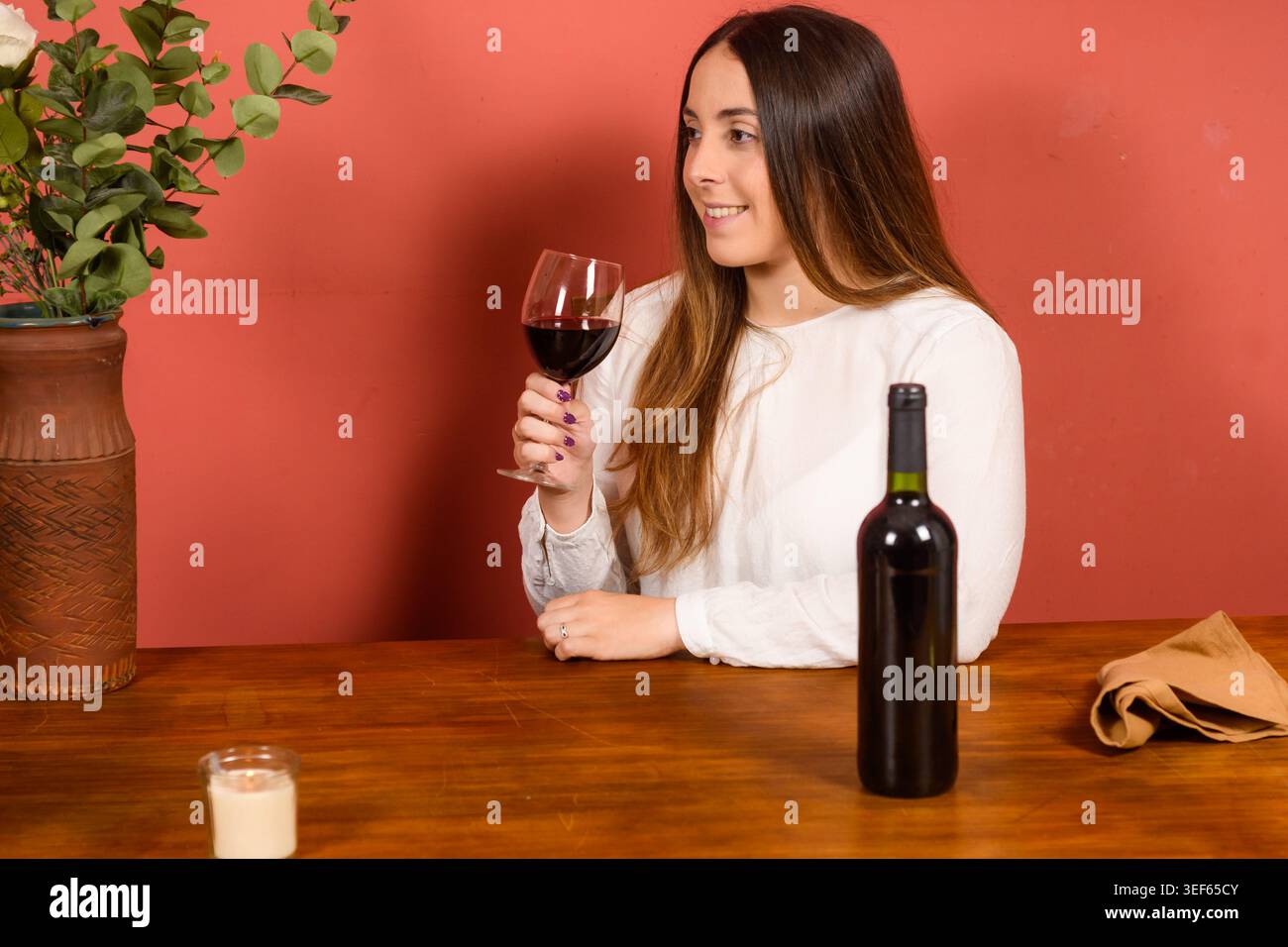 Junge Frau trinkt Rotwein lächelnd, entspannter Lebensstil und Genusskonzept in warmer ästhetischer Umgebung Stockfoto