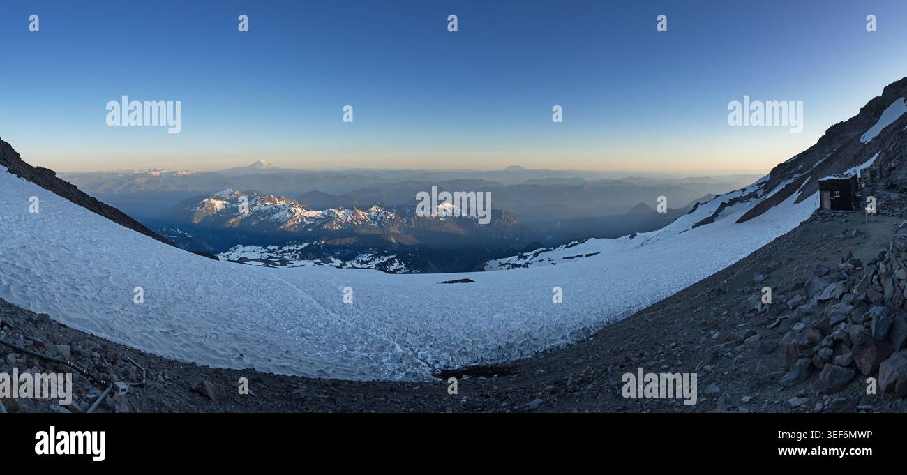 Panorama mit Blick auf das Camp Muir auf dem Mount Rainier mit entfernten vulkanischen Bergen am Horizont und der Tatoosh Range beleuchtet von der untergehenden Sonne Stockfoto