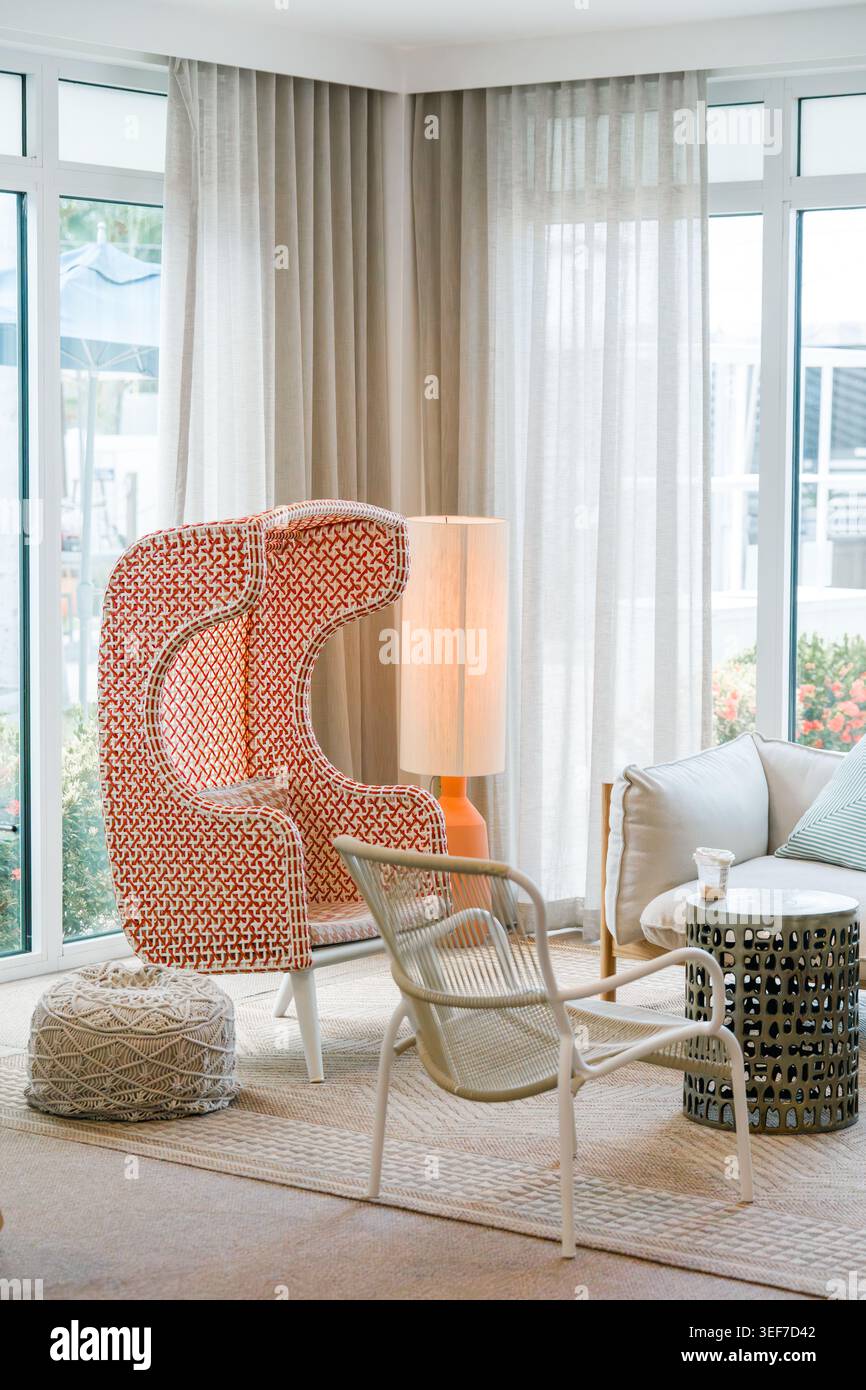Ein heller, moderner Lounge-Bereich verfügt über einen einzigartigen orange-weiß gemusterten Flügelstuhl mit hoher Rückenlehne, der sich in der Nähe eines großen Fensters befindet. Stockfoto