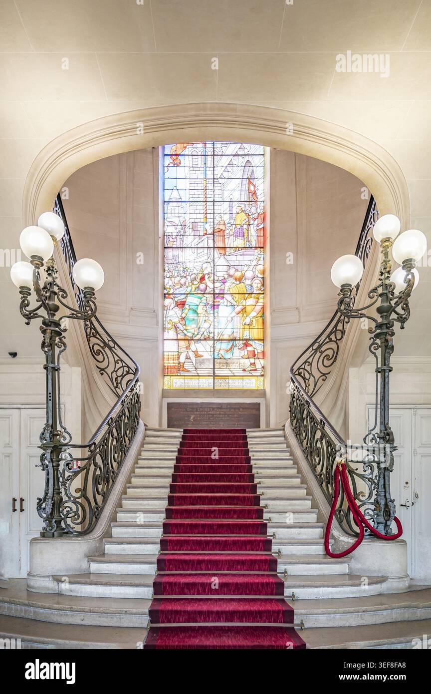 Herrliche Treppe mit rotem Teppich, schmiedeeisernem Geländer und historischem Kandelaber vor einem bunten Buntglasfenster im Hotel de Ville Stockfoto