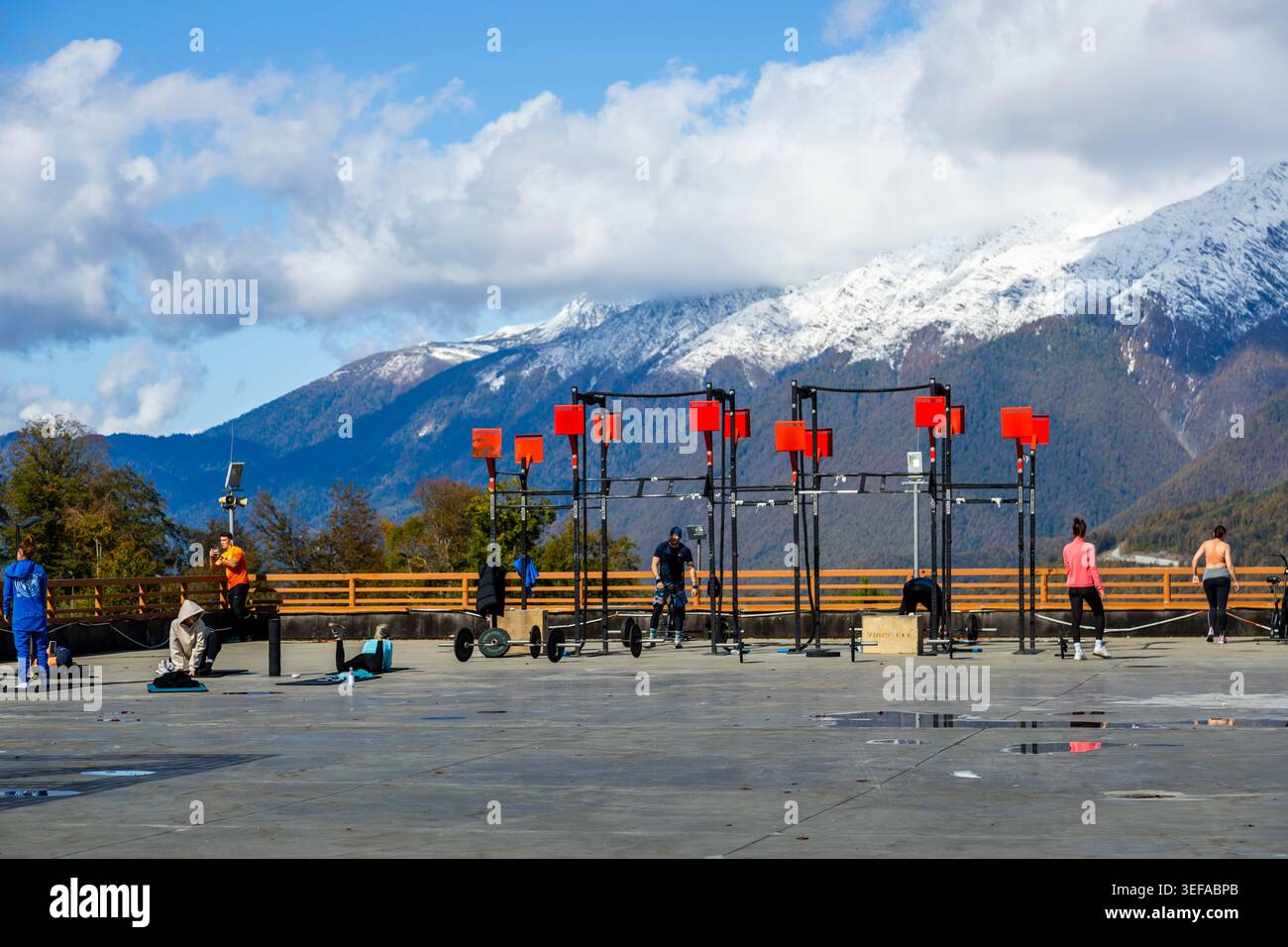 Gruppen von Personen heben Gewichte an und nutzen Fitnessgeräte im Freien. Schneebedeckte Berge und blauer Himmel bilden die Kulisse. Es ist tagsüber und die Gegend ist gut gepflegt Stockfoto