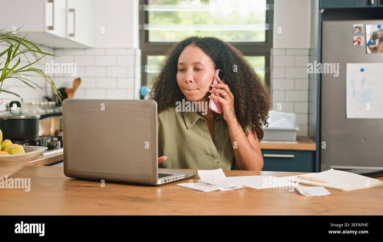 Junge Frau, Die Von Zu Hause Aus Am Küchentisch Auf Laptop Arbeitet, Während Sie Telefoniert Und Rechnungen Überprüft Stockfoto