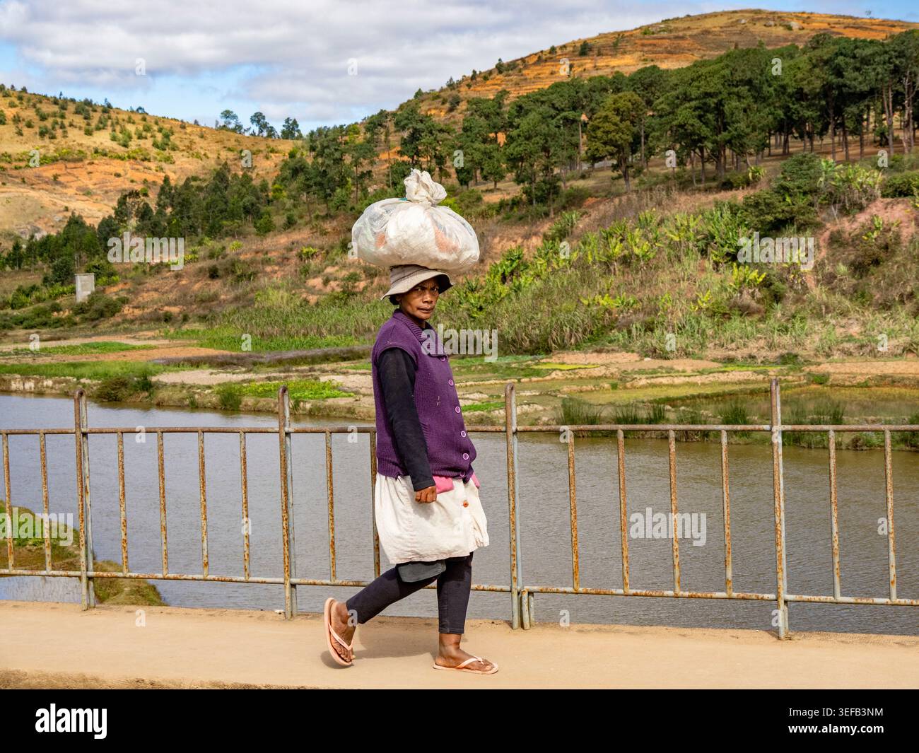 Szenen entlang der Autobahn zeigen eine Person, die mit einer Last auf dem Kopf in der Nähe eines Flusses in Madagaskar läuft Stockfoto