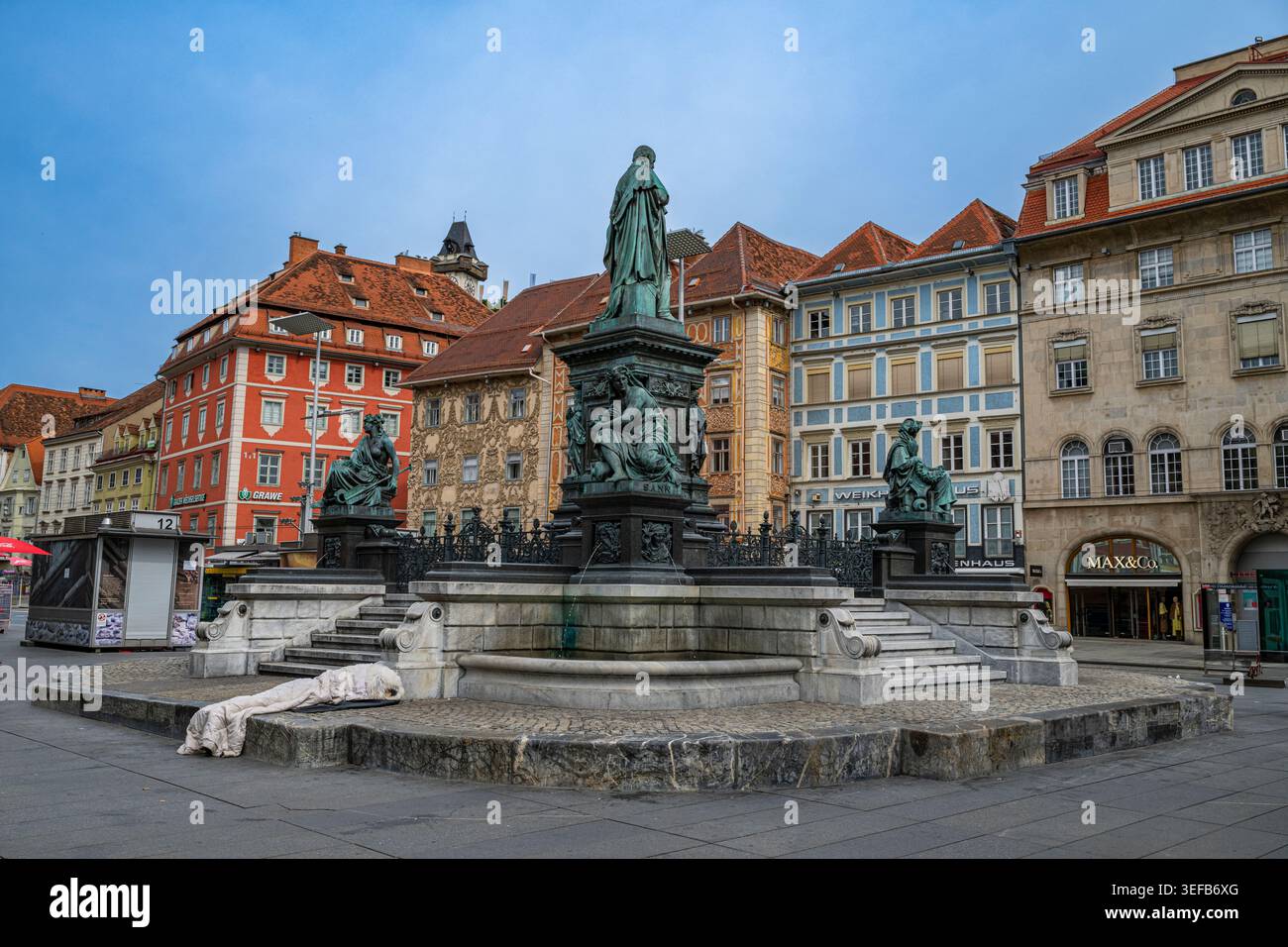 Brunnen in der historischen Gegend von Graz mit umliegenden Gebäuden und bewölktem Himmel Stockfoto
