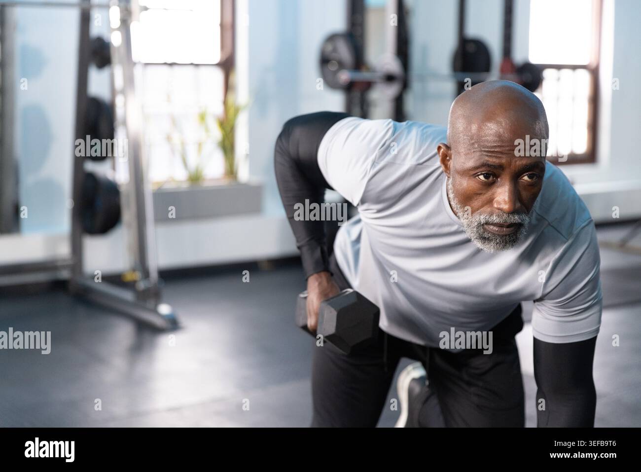 Ein ranghoher Afroamerikaner, der eine Reihe im Fitnessstudio führt, eine Hexenhantel hält und Sportbekleidung trägt. Fitness, Gewichte, Training, Kraft, Sonnenlicht, stu Stockfoto