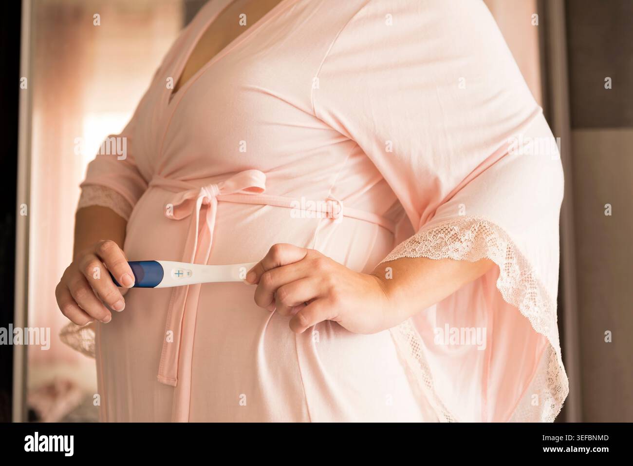 Schwangere Frau, die einen positiven Schwangerschaftstest hält, während sie sanft ihren Babybummel berührt und einen zärtlichen und emotionalen Mutterschaftsmoment festhält. Weich, natürlich Stockfoto