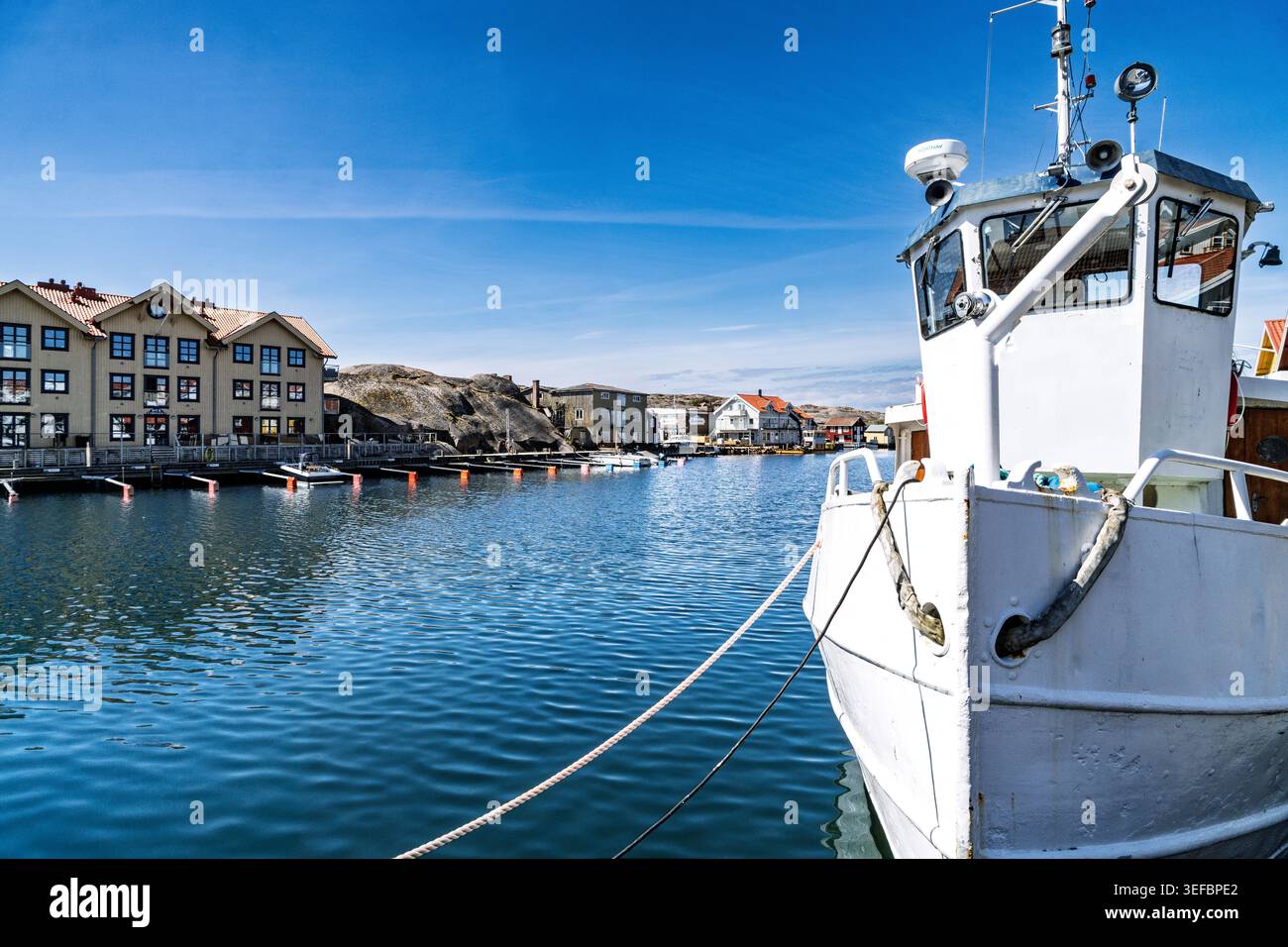 Das Boot ist am Ufer mit einem Seil am Dock verankert. Farbenfrohe Häuser und Gebäude säumen den Hafen in einer kleinen Küstenstadt. Stockfoto